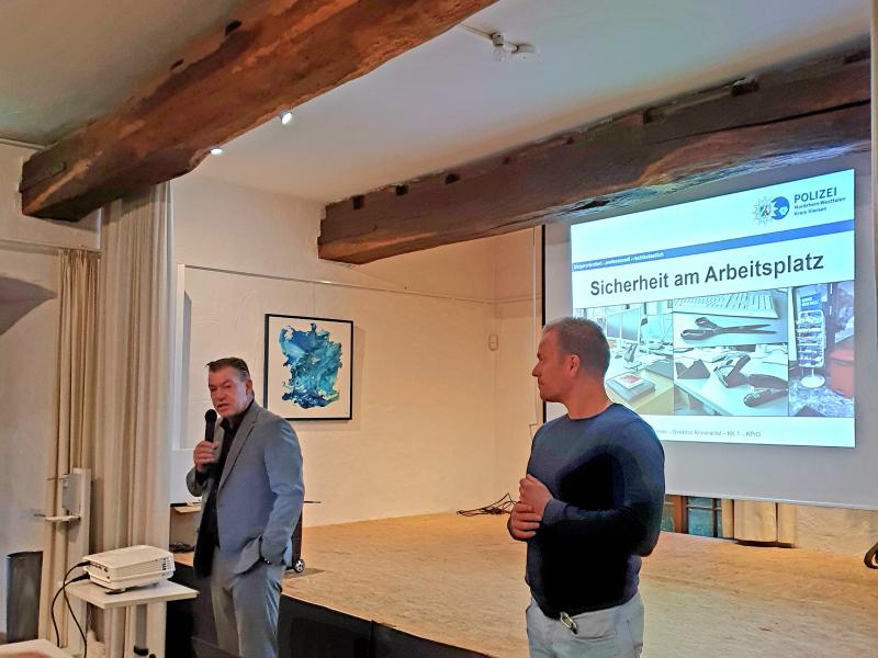 Bürgermeister Gellen links in grauen Anzug und schwarzen Hemd hält Mikrofon in rechten Hand und linke Hand in Hosentasche, rechts blonder Mann mit Pullover und heller Jeans, vor ihnen Tisch mit Stühlen, iPad, Laptop und Trinkflaschen sowie weißer Beamer, beide stehen vor Bühne mit weißer Leinwand und Präsentation zu „Sicherheit am Arbeitsplatz“, im Hintergrund weiße Wand mit Bild und weißer Vorhang, weiße Decke mit Holzbalken und Lampen