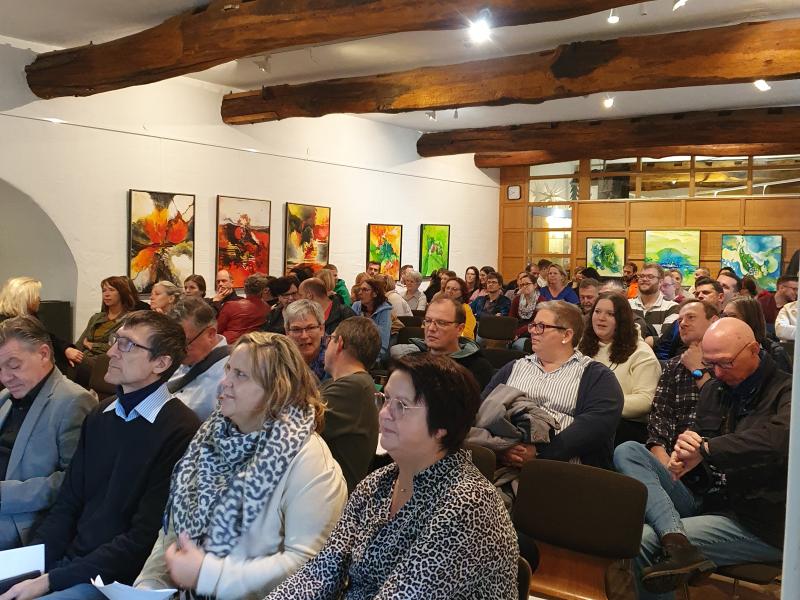 Personen sitzen auf dunklen Stühlen in Reihen hintereinander, Holzboden, im Hintergrund weiße Wand und Holzwand mit Scheiben und Bildern, weiße Decke mit Holzbalken und Lampen