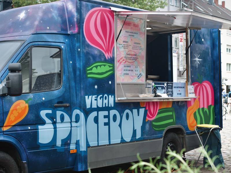 Ein blauer Foodtruck steht auf einem asphaltierten Platz. Eine Luke mit Abdeckung ist offen und eine Speisekarte und ein Tresen ist zu sehen. Auf dem Fahrzeug selber ist der Schriftzug Vegan Spaceboy drauf lackiert mit vielen bunten Gemüsesorten. Im Vordergrund und Hintergrund befinden sich grüne Pflanzen.