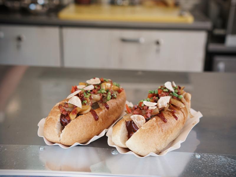 Zwei mit Gemüse gefüllten länglichen Brötchen stehen auf einer silbernen Ablagefläche bereit zum Servieren. Neben Gemüse sind auch Toppings auf dem Brötchen gestreut wurden und eine geschwungene Linie aus Soße ziert die Speise.