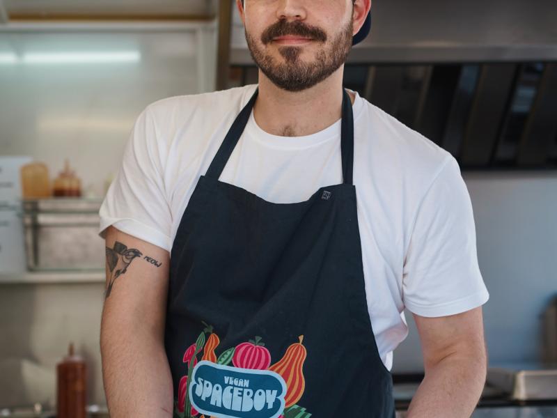 Auf dem Bild ist ein Herr im mittleren Alter zu sehen. Er hat ein weißes T-Shirt an und trägt eine schwarze Schürze mit einem pink blauen Aufdruck "Vegan Spaceboy". In der Hand hält er vegane Imbissgerichte. Im Hintergrund ist ein Tresen zu sehen und das Innere eines Foodtrucks.
